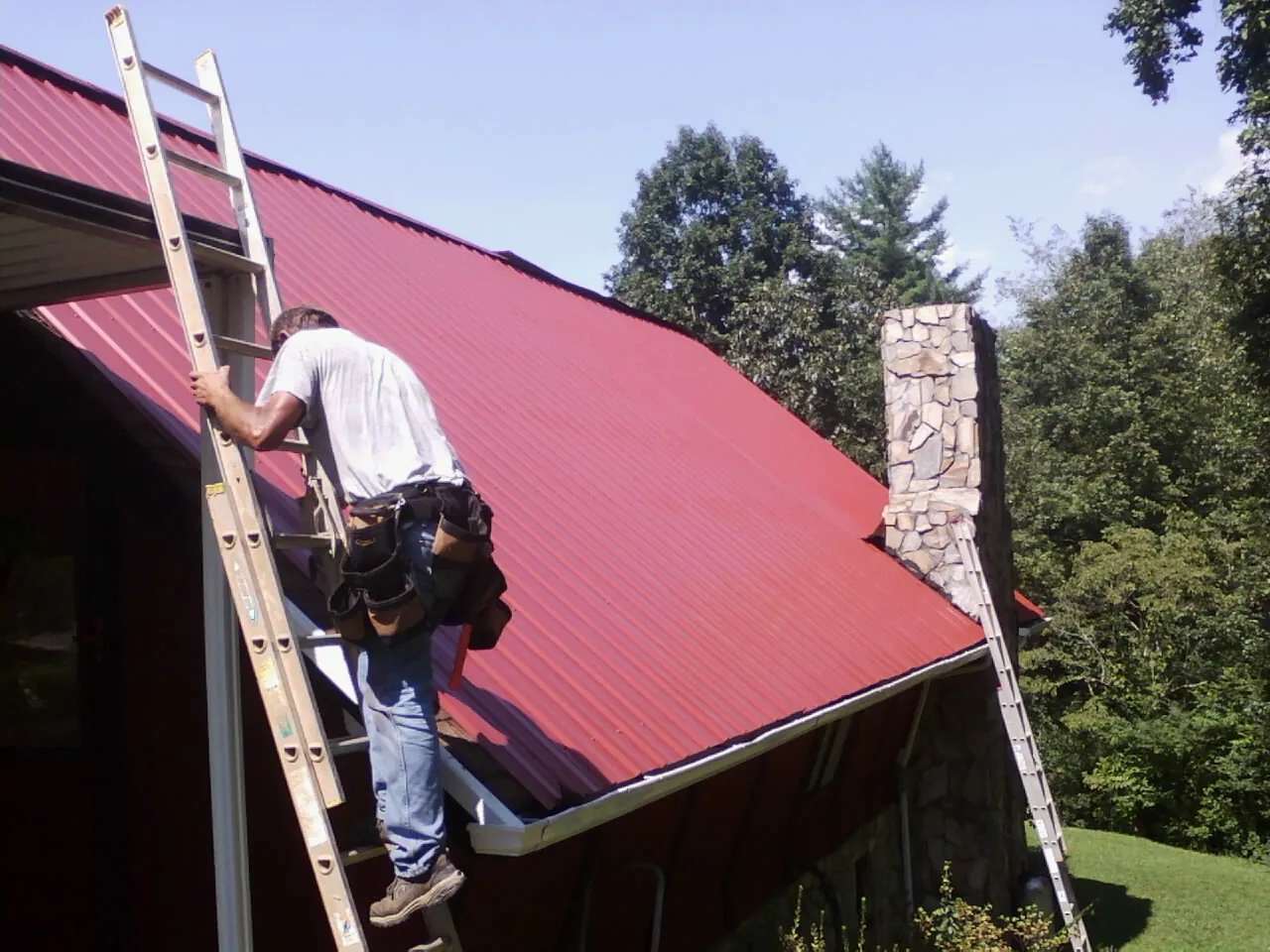 Roofer on a ladder beside a red metal roof and stone chimney — Soffit & Fascia Repair by Roofing Brothers in Spencer